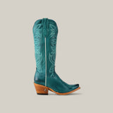 The Kellie Turquoise Leather Sole boot, featuring a tall shaft and snip toe, showcases intricate stitching. Its wooden stacked heel enhances the polished leathers vibrant look against a light beige background.