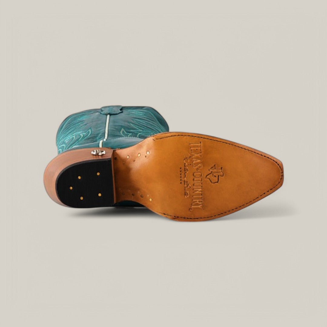 A turquoise Kellie leather boot with a snip toe is laid on its side, displaying its tan sole marked with the TEXAS COUNTRY logo and decorative stitching, while intricate lighter embroidery adorns its tall shaft.