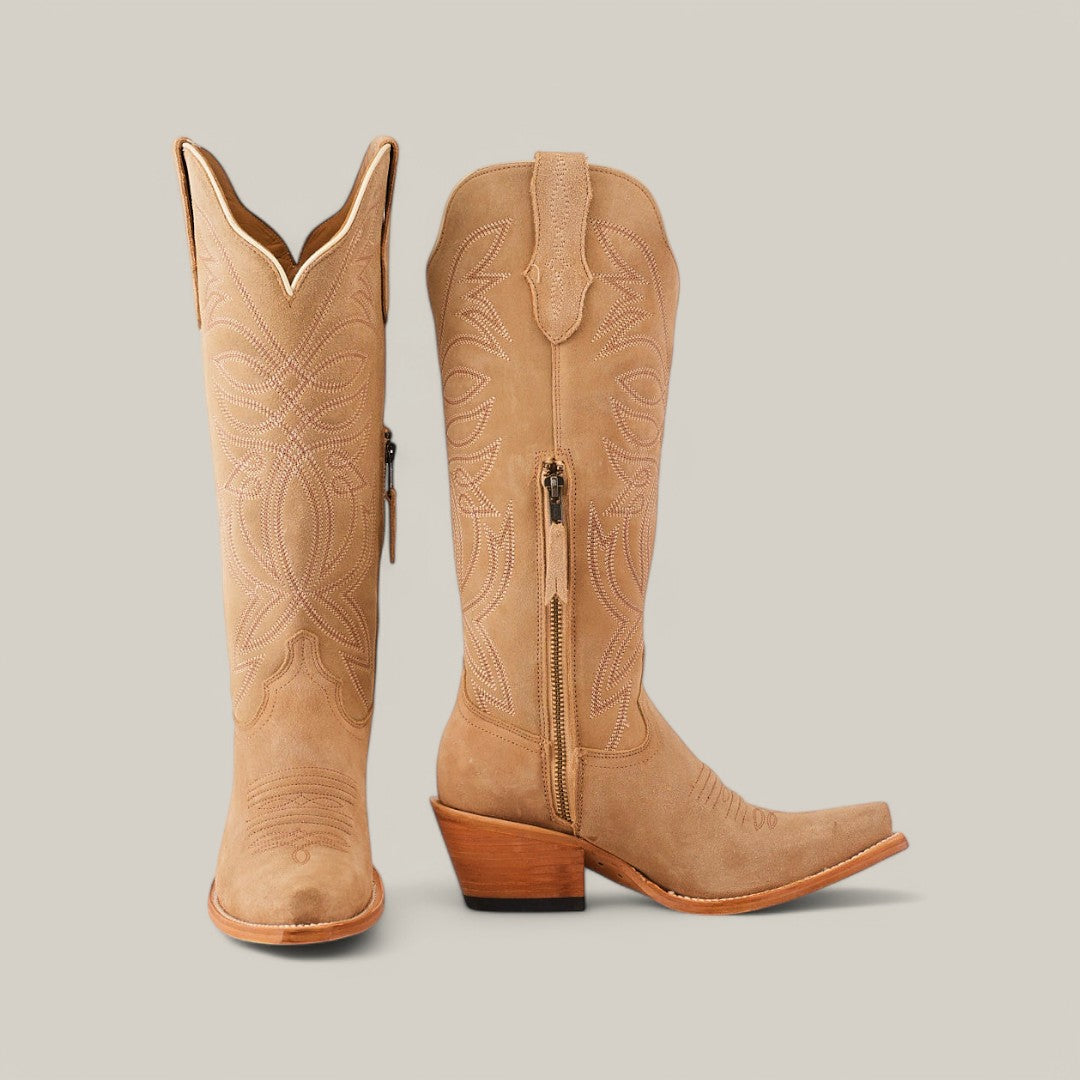 The Kellie Suede Fawn boots, crafted with a premium leather sole, are displayed against a neutral background. One boot stands upright showcasing the decorative stitching and tall shaft, while the other reveals its snip toe design and wooden heel. A side zipper enhances their elegance.