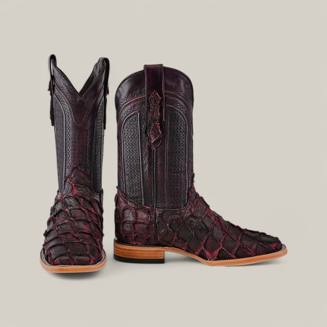 A pair of intricately designed Exotic Pirarucu Fish cowboy boots in Matte Black Cherry features a square-toe silhouette and detailed stitching. One boot is upright, while the other tilts, highlighting the textured scales. The soles are made from light brown leather.