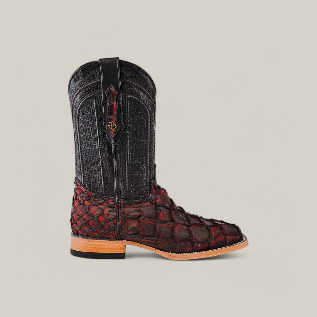 The Exotic Pirarucu Fish - Red - Square Toe boot showcases a dark upper with intricate stitching and a textured, scaled red lower section. Set against a light neutral background, this unique cowboy boot features a light tan heel and sole, embodying elegance and distinctiveness.