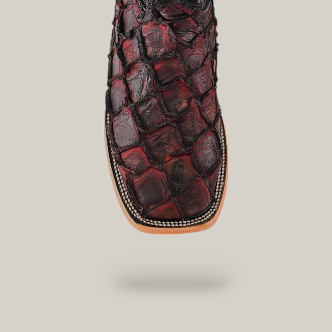 A close-up of the Exotic Pirarucu Fish boot in dark red and black, showcasing a textured, scale-like design from exotic leather. It features a square toe with intricate stitching and a light-colored sole. The plain light gray backdrop enhances its striking appeal.