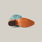 The image features the sole of an Olimpia Green Teel - Tall Shaft - Snip Toe cowboy boot with intricate patterns on the tan base. Tilted on its side, it showcases a turquoise upper adorned with decorative stitching against a plain, neutral background.