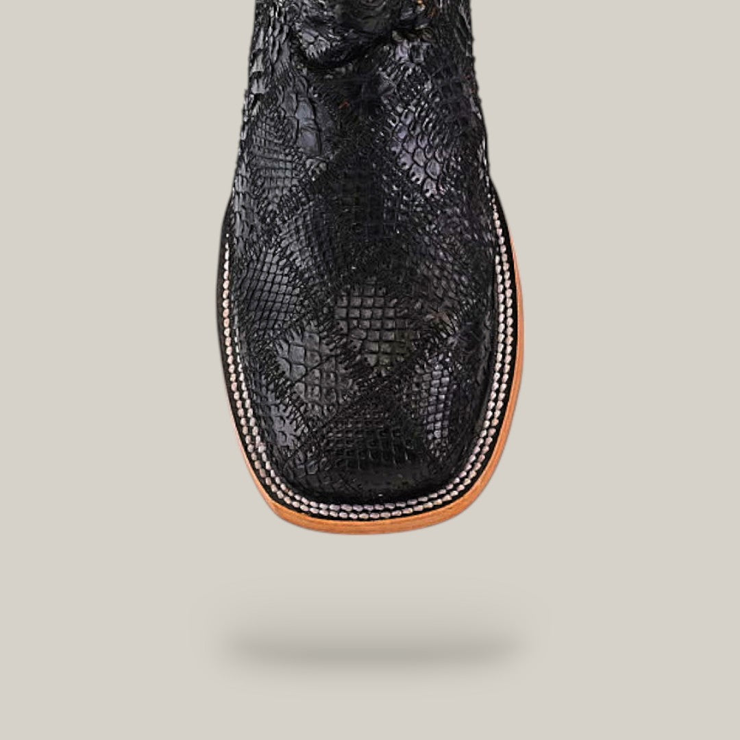 A close-up top view of the Exotic Patchwork Python - Black - Square Toe boot showcases its textured snakeskin pattern, black square-toed design, detailed white stitching on a light brown welt, set against a beige background.