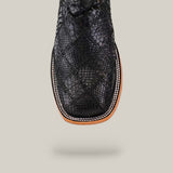 A close-up top view of the Exotic Patchwork Python - Black - Square Toe boot showcases its textured snakeskin pattern, black square-toed design, detailed white stitching on a light brown welt, set against a beige background.