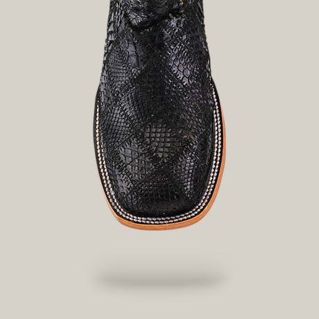 A close-up top view of the Exotic Patchwork Python - Black - Square Toe boot showcases its textured snakeskin pattern, black square-toed design, detailed white stitching on a light brown welt, set against a beige background.