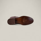 The Wyatt - Cogñac - Roper Toe is a brown leather dress shoe, shown from the bottom with a smooth, polished sole, small metal nails at the heel, and embossed branding near the arch. Set against a light beige background.