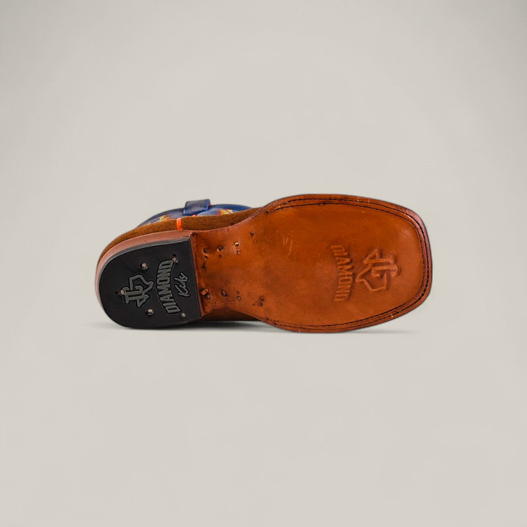 A close-up of the Diamond Suede Ocre - Square Toe - Kids shoe sole shows brown leather with stitched edges, a black rubber heel cap, and embossed logos on both the sole and heel. The background is plain and light-colored.