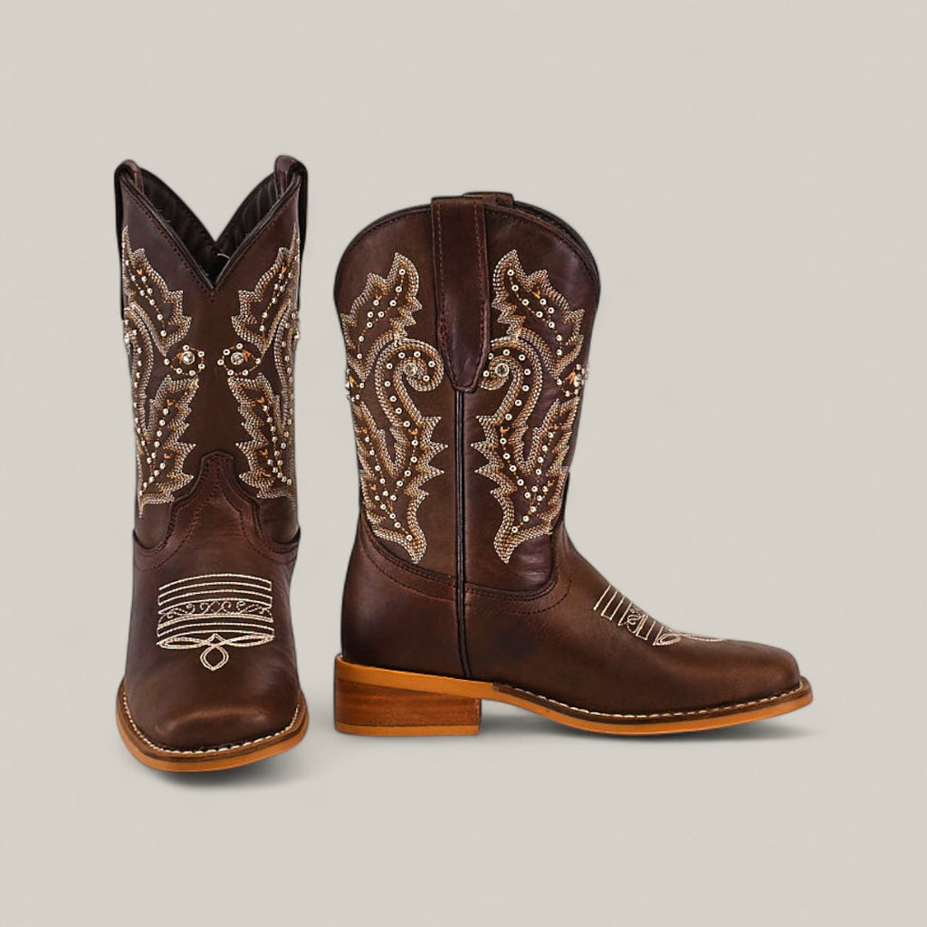 The Britany Brown Square Toe Kids Boots, made from quality leather with beige embroidery and stud details, are displayed with one boot facing forward and the other in profile to showcase their craftsmanship.
