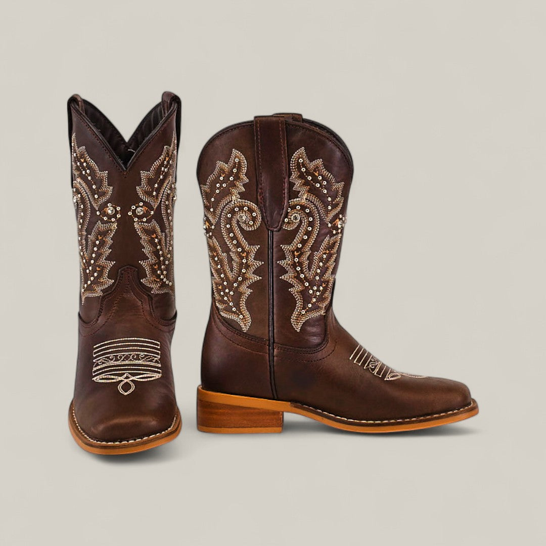 The Britany Brown Square Toe Kids Boots, made from quality leather with beige embroidery and stud details, are displayed with one boot facing forward and the other in profile to showcase their craftsmanship.