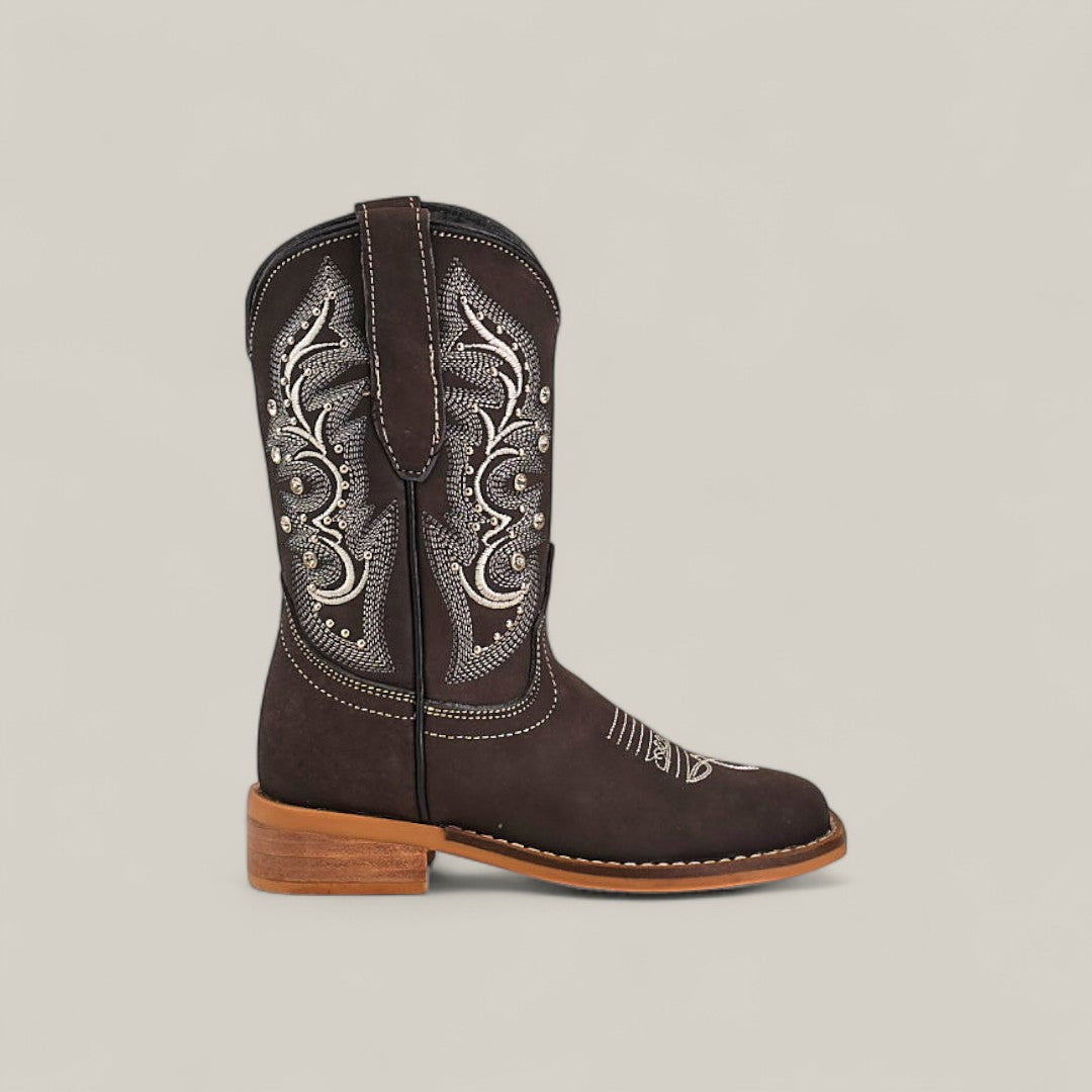 The Amara Suede Oxford - Square Toe Kids Boots are shown in profile, featuring dark brown suede, white embroidery, silver studs on the shaft, pull straps, and a light brown stacked heel and sole against a neutral background.