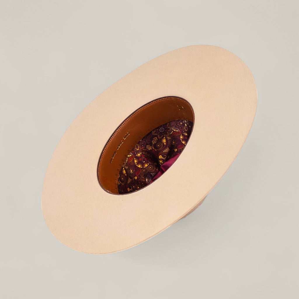 The 10x Belly - TC Hats features a wide brim, light beige Western style, viewed from above with a brown leather inner band and vibrant paisley-patterned fabric lining inside its shapeable crown.