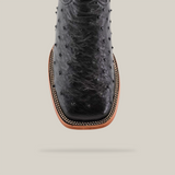 A close-up of the Exotic Full Quilt Ostrich - Black - Square Toe boot highlights its dotted texture, wooden sole, and intricate stitching against a plain light gray background.