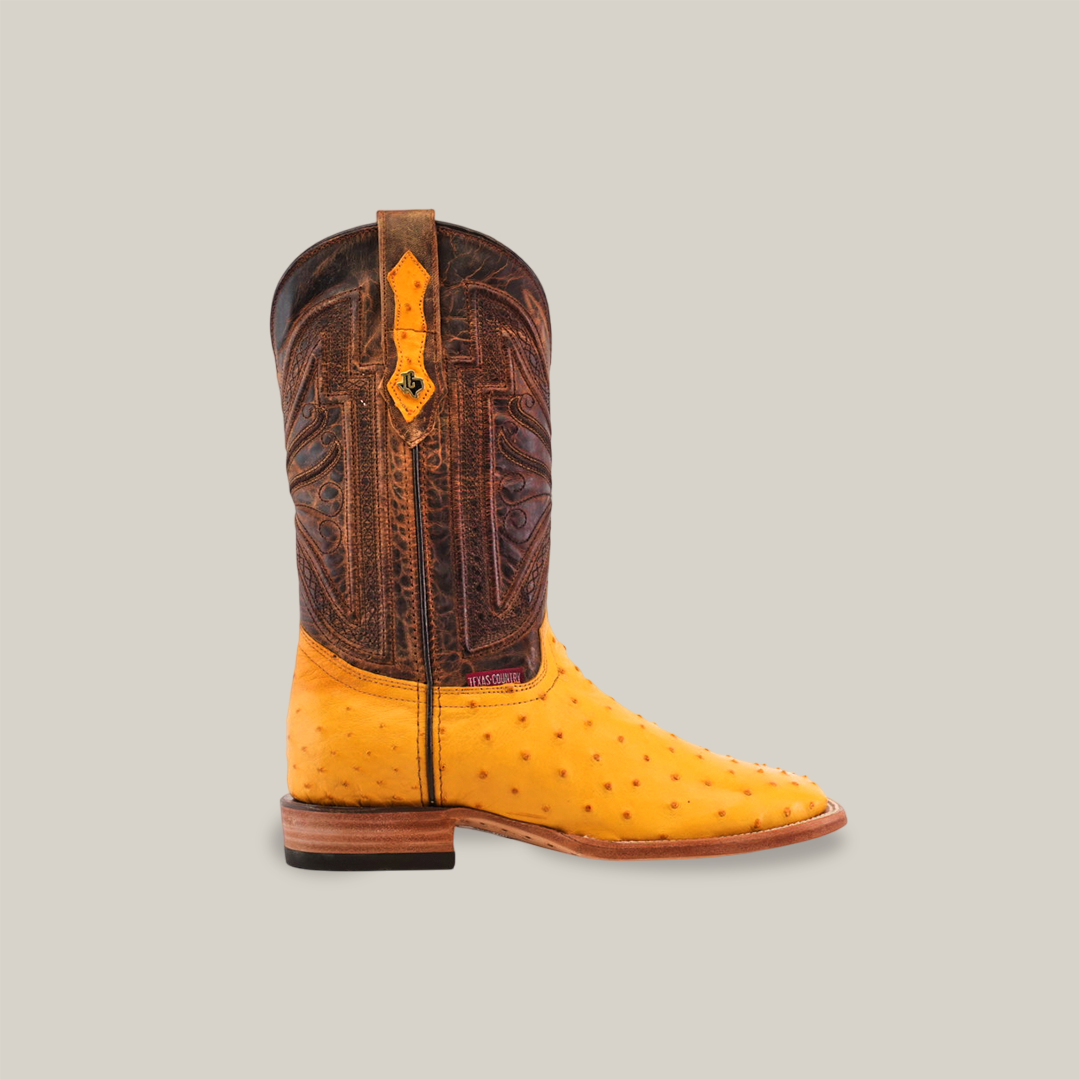 The Exotic Full Quilt Ostrich - Buttercup- Square Toe boot features a buttercup-colored ostrich leather foot with a dark brown, intricately patterned shaft, complemented by a wooden heel and decorative pull strap, all set against a plain beige background.