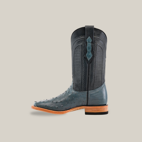 The Exotic Full Quilt Ostrich - Blue Jean boot stands out with a sleek square toe against a plain background. Crafted from textured dark leather like ostrich skin, it features decorative stitching, a wooden heel and sole, and a high shaft with pull straps—ideal for any Western wardrobe.