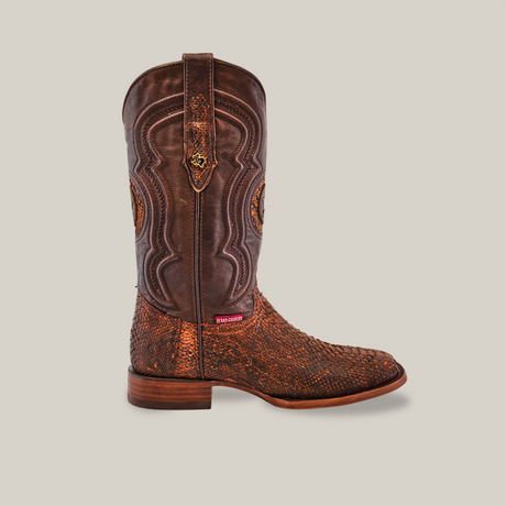 The Exotic Python cowboy boot in rustic cogñac features ornate stitching, detailed texture, and a square toe. It has a wooden heel, toe accents, and a red tag by the sole against a plain beige background.