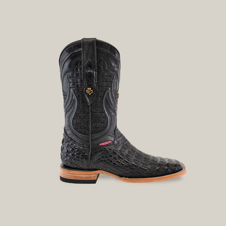 The Exotic Caiman Horn Back cowboy boot, in black with a square toe, features luxurious caiman leather with crocodile texture, decorative stitching on the shaft, a brown sole and heel, and a small side label. Displayed against a light beige background.