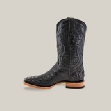 The Exotic Caiman Horn Back Black boot features intricate stitching and an embossed pattern in luxurious caiman leather. This handcrafted boot showcases detailed craftsmanship with a square toe, low stacked heel, and tan leather sole against a plain beige background.