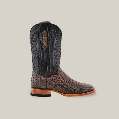 A close-up of the Exotic Caiman Horn Back - Serpentine boot, showcasing its black leather upper and textured brown lower half with detailed stitching. It features a wooden heel and a square toe, set against a plain beige background.