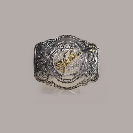 The Floral Bull Rider Buckle, crafted from alpaca silver, showcases intricate western designs and a raised gold-colored bull rider in the center. Its elegantly presented on a clear stand against a plain gray background.