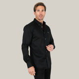 A man in a Men’s Single Pocket Logo Modern Fit Stretch Dress Shirt (Black, CTL9258) stands against a light background. The stretch fabric of the modern fit enhances his silhouette. He has short hair and gazes slightly to the left.