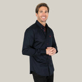 A smiling man wearing the Men’s Single Pocket Logo Modern Fit Stretch Dress Shirt in navy, featuring striking red buttons, stands against a beige background. The stretch fabric ensures comfort as he clasps his hands in front.