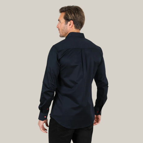 A man with brown hair stands facing away, wearing a CTL9262 Men’s Single Pocket Logo Modern Fit Stretch Dress Shirt in navy and black pants. The stretch fabric offers comfort and style against the solid light gray background.