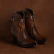 The CU483 Maple Embroidery & Harness - Round Toe boots are crafted from premium brown leather with intricate maple embroidery and decorative stitching. They feature a buckle strap and stacked heels, set against a matching brown background.