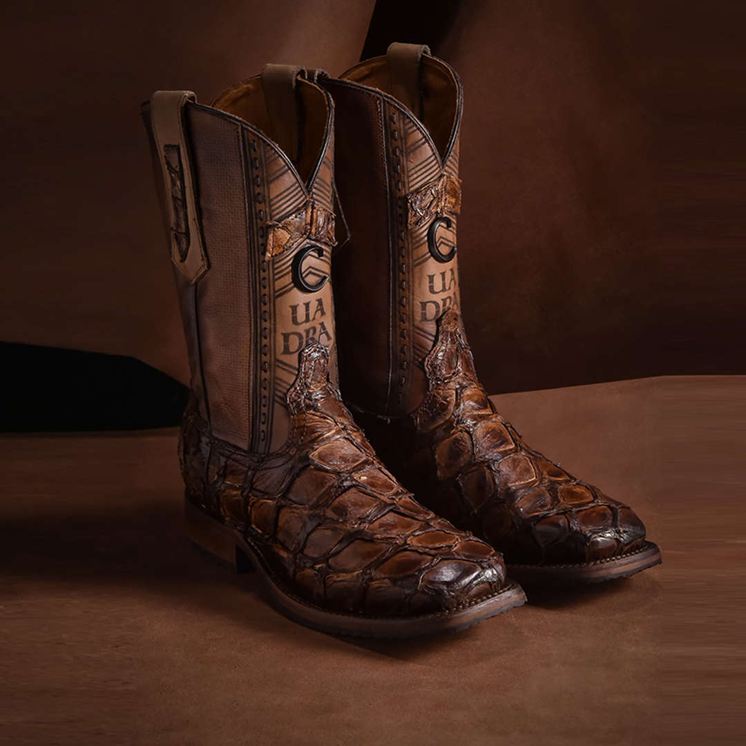 Chocolate Glazed Fish Pirarucu Laser & Woven Square Toe boots made of brown leather with an alligator skin texture stand upright on a matching brown surface. Featuring detailed stitching and a UA DRA logo on the shaft, these are luxury cowboy boots.