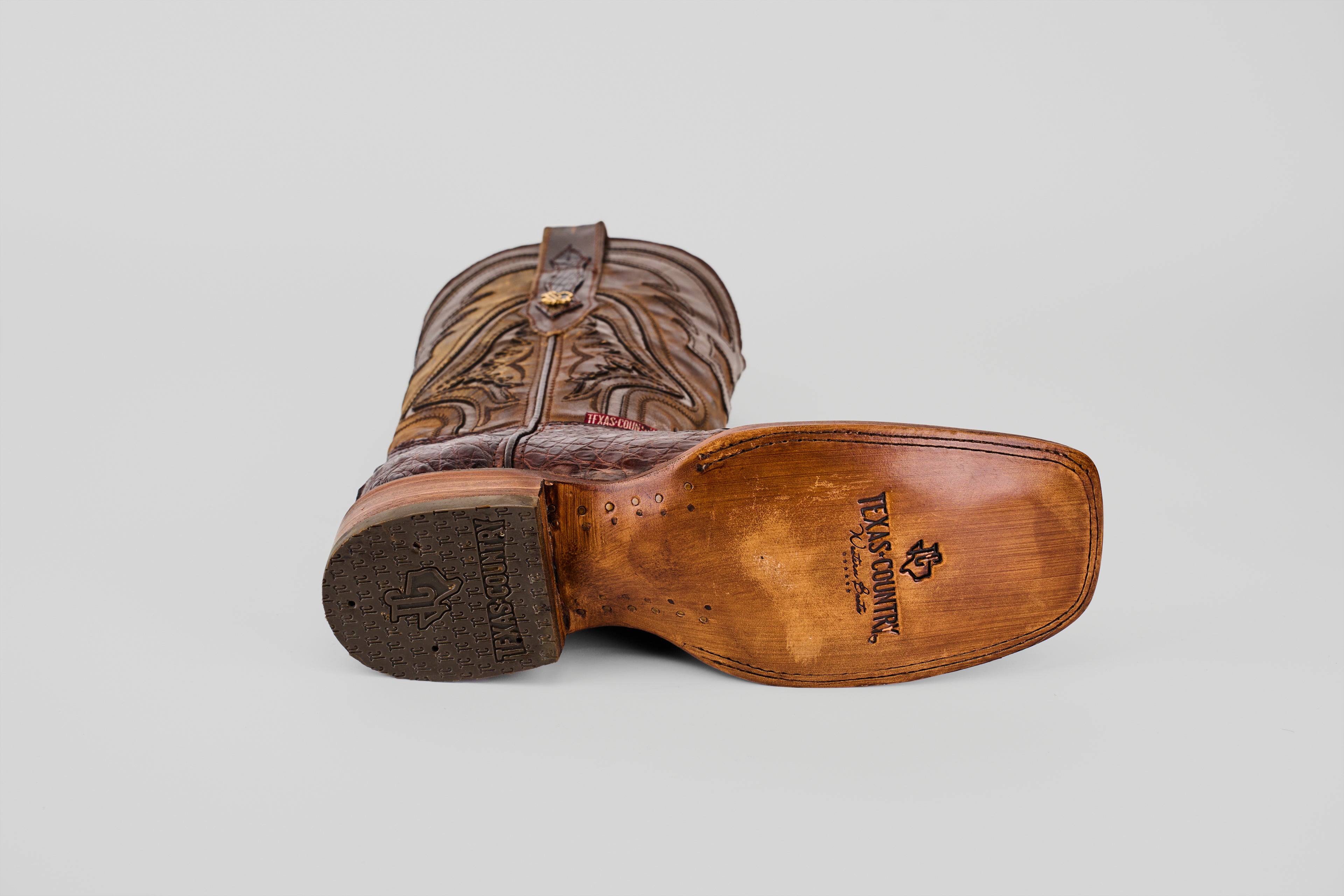 A brown Exotic Caiman Hornback - Brown - Rodeo Toe boot rests on its side, displaying the Texas Country logo stamped on genuine caiman leather, highlighting its premium quality against a plain gray background.
