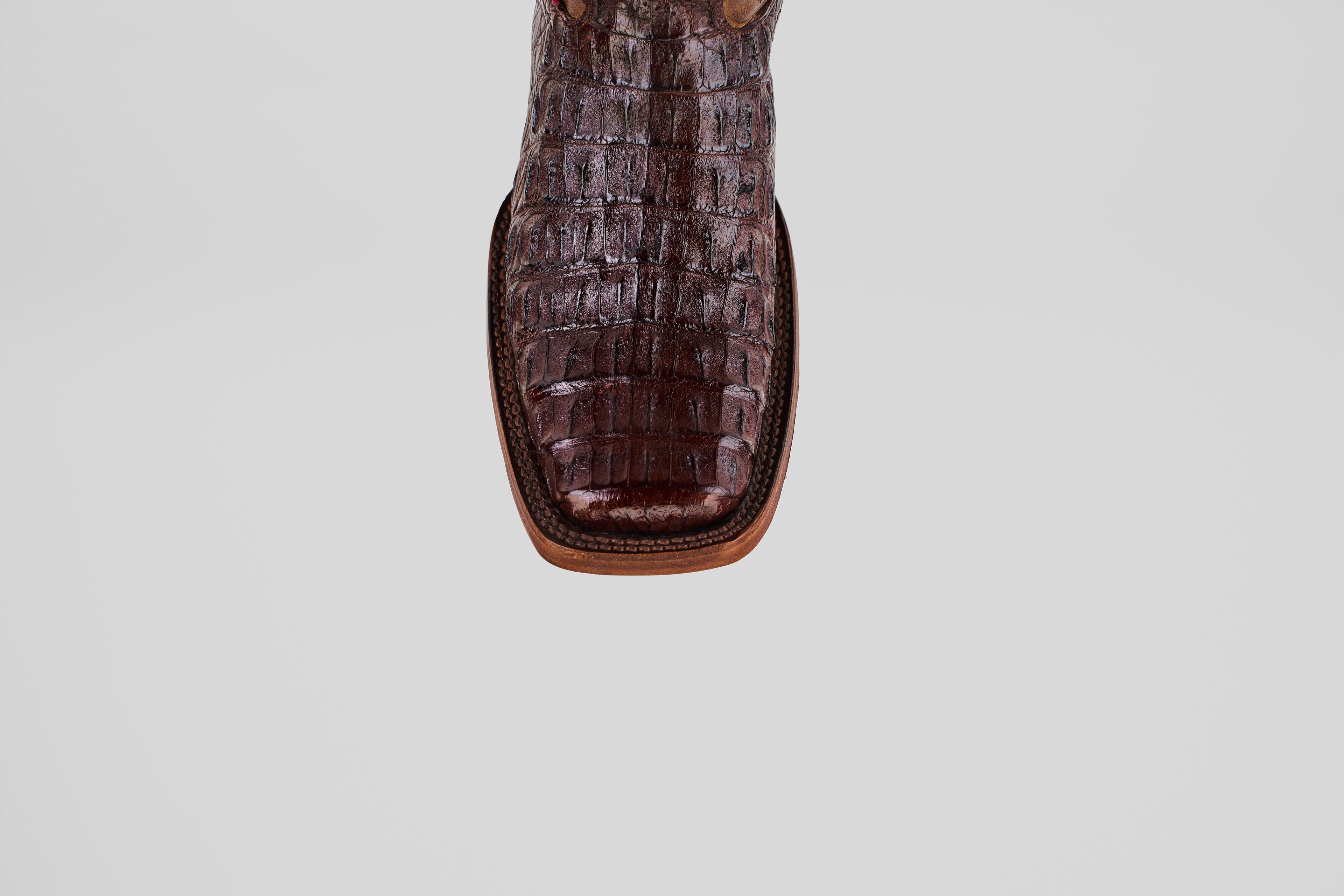 A close-up, top-down view showcases the unique texture of the Exotic Caiman Hornback - Brown - Rodeo Toe boots square toe in genuine caiman leather against a plain light gray background.
