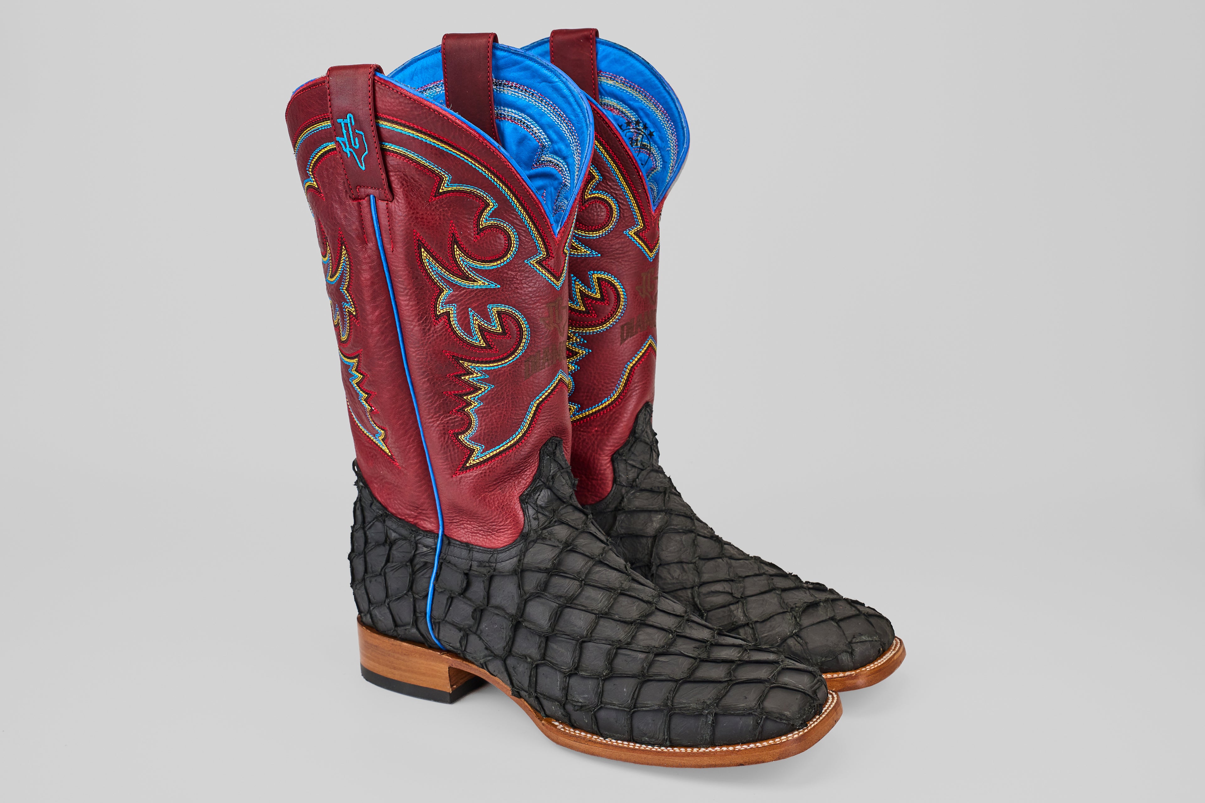 The Dodge - Matte Black - Square Toe boots feature red and blue decorative uppers, textured black Pirarucu leather feet, wooden heels, and light blue stitching accents, displayed against a plain gray background.