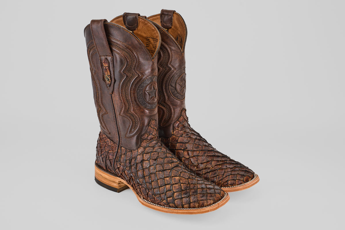 A pair of Exotic Pirarucu Fish - Brown - Square Toe boots with textured, patterned leather and decorative shaft stitching, displayed on a light gray background.