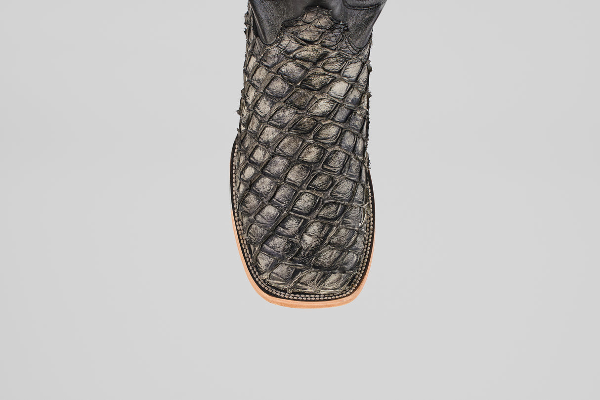 A close-up of the Exotic Pirarucu Fish - Newspaper - Square Toe boot showcases its black, scale-patterned exotic leather and brown sole, set against a plain light gray background.