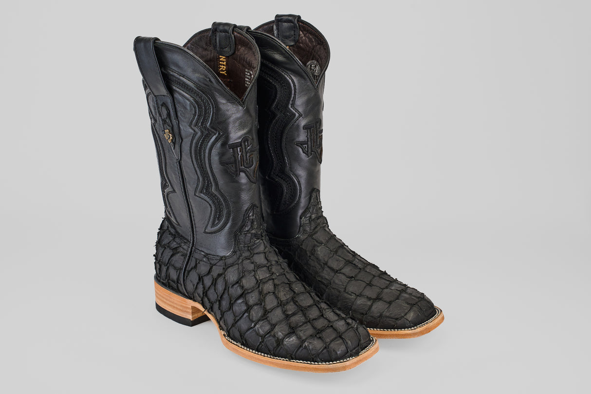 A pair of Exotic Pirarucu Fish - Matte Black - Square Toe boots featuring a scale-like texture, smooth leather shafts, light brown wooden soles, and low heels.