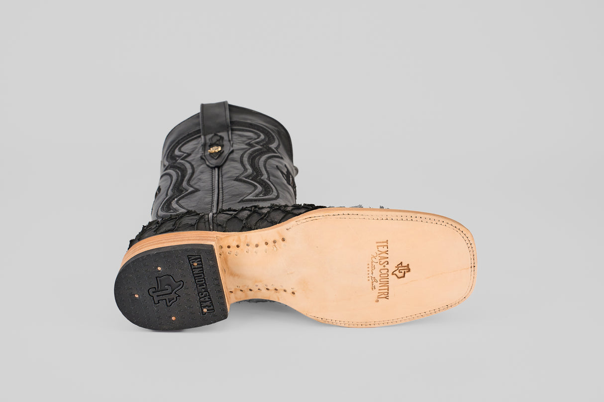 A black Exotic Pirarucu Fish boot with a square toe lies on its side, highlighting intricate stitching and the Texas Country logo on the sole—a striking mix of matte black textures and premium exotic leather.
