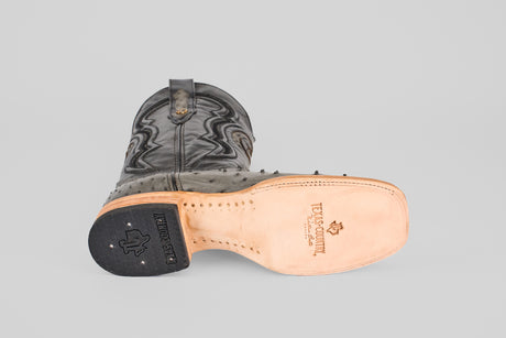 A single Exotic Full Quilt Ostrich - Gray - Square Toe boot lies on its side, showing its tan leather sole with visible stitching, Texas Country logo, black rubber heel, and classic ostrich-inspired decorative stitching on the upper.