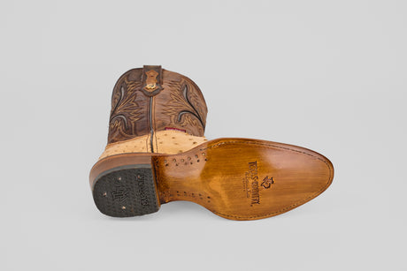 The Exotic Full Quilt Ostrich - Orix - Round Toe boot lies on its side, revealing a leather sole with stitching and a brand logo. Its brown upper features decorative stitching and a textured beige ostrich section near the ankle for classic Western style.