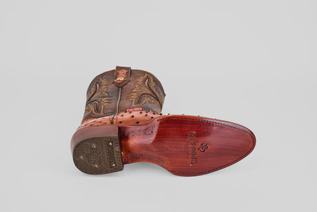 A single Exotic Full Quilt Ostrich - Brandy - Round Toe cowboy boot rests on its side, highlighting intricate stitching and a polished wooden sole with the Tecovas logo against a plain light gray background.