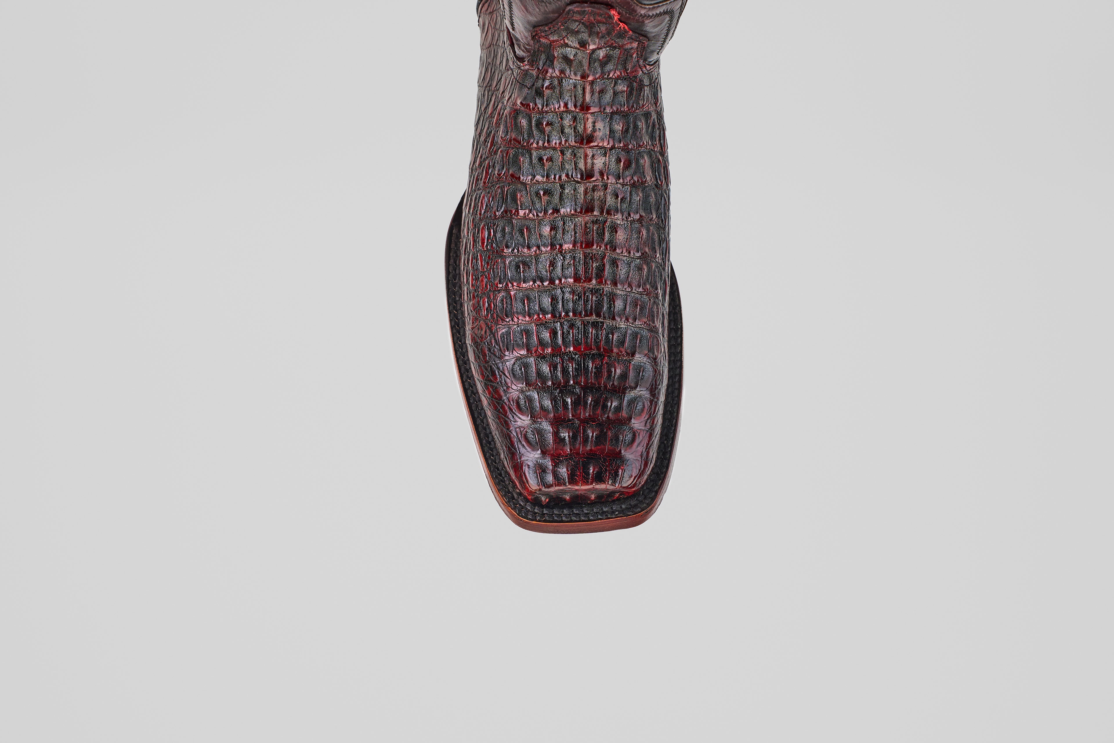 A close-up top view of the Exotic Caiman Hornback - Blackcherry boot highlights its Rodeo Toe and crocodile-patterned leather, photographed against a plain gray background—ideal for fans of this exotic style.