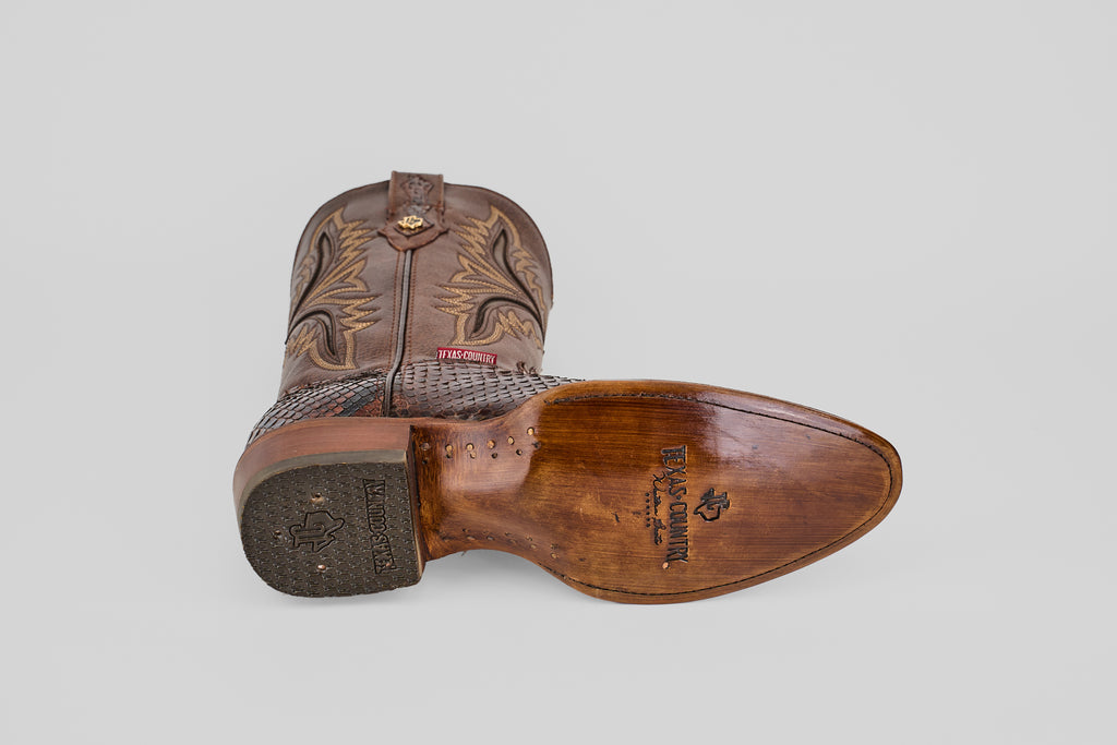 The Tanner - Brown - Round Toe cowboy boot is shown on its side, featuring intricate shaft stitching and a textured, partially worn sole with the Texas Country logo and Python Cafe Oval imprint.