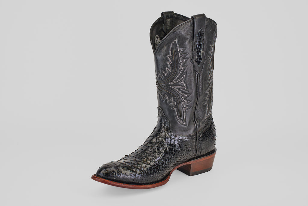 The Tanner - Black - Round Toe is a single black leather cowboy boot with intricate shaft stitching and a Python Negro Oval patterned foot, featuring a brown stacked heel and sole, displayed on a plain light gray background.