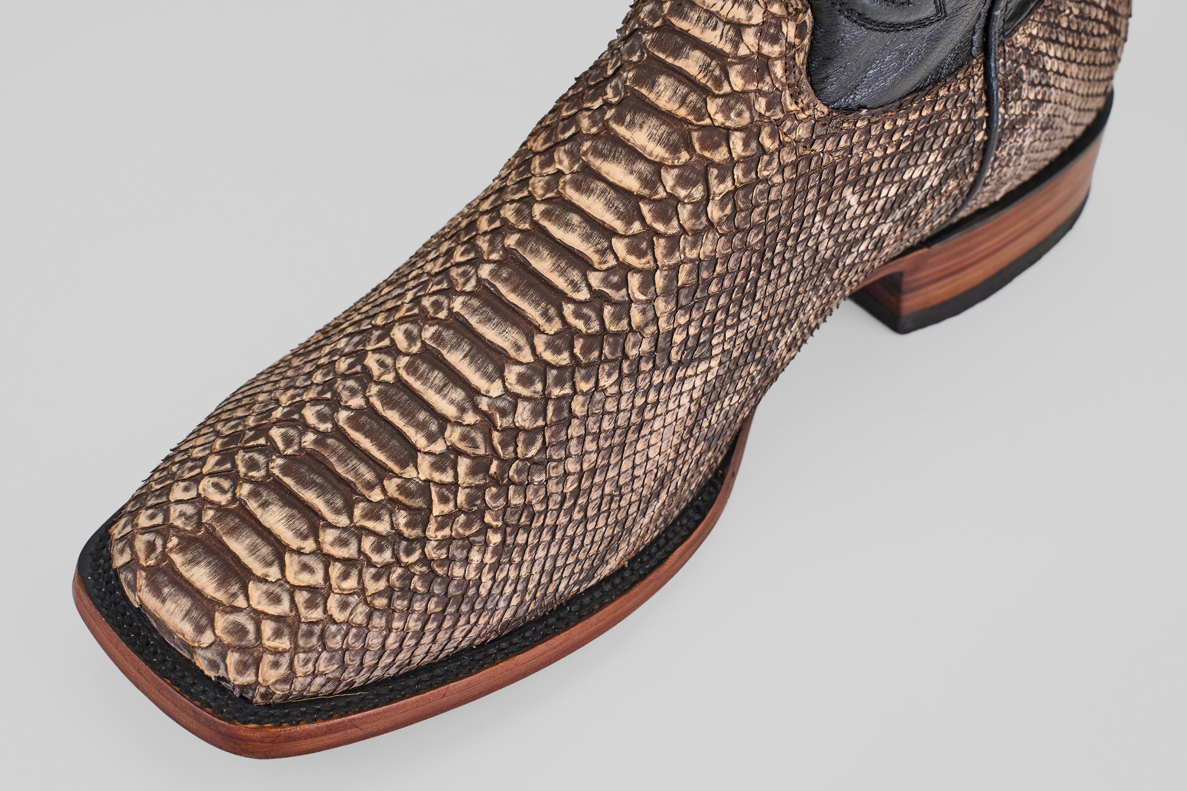 A close-up of the Tanner - Rustic Bone - Square Toe boot highlights its brown python design, black trim, and wooden sole, showcasing Western craftsmanship against a light gray background.