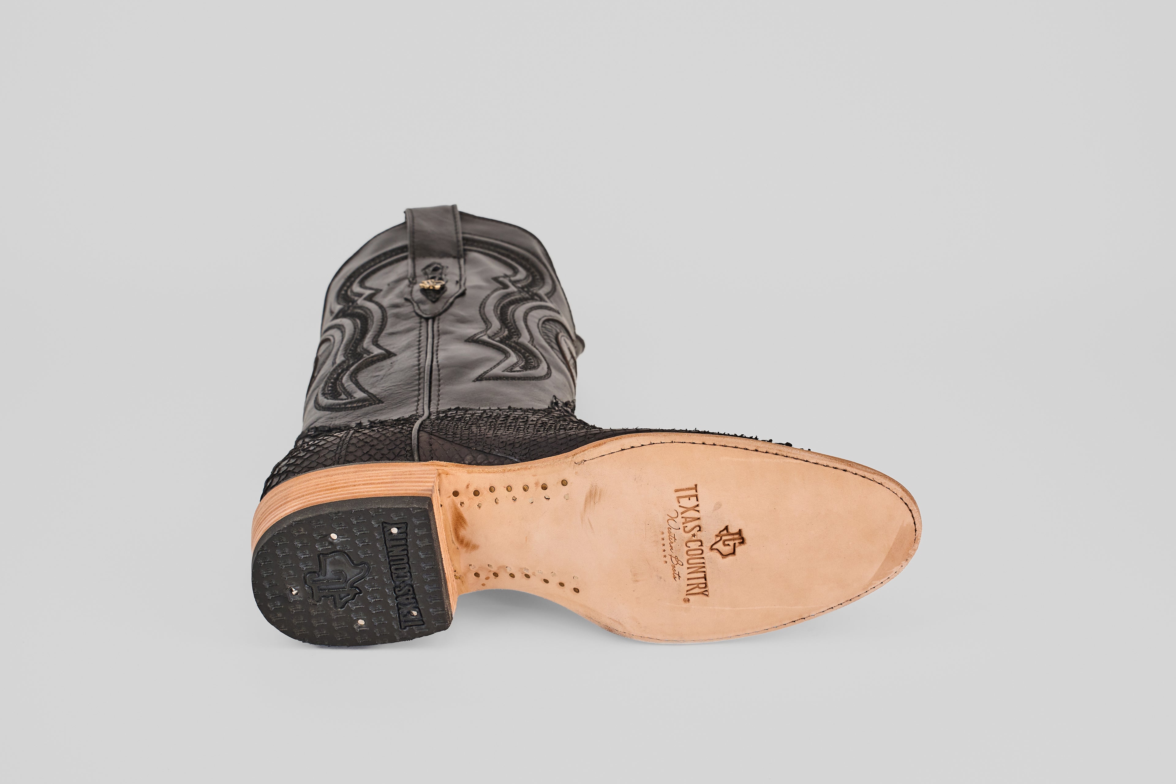 The Tanner – Matte Black – Roper Toe boot is shown on its side, highlighting the “Texas Country” stamped leather sole. This western-inspired boot features textured matte black leather and decorative stitching on the shaft.