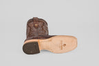 The Tanner - Brown - Square Toe boot lies on its side, displaying decorative stitching on the upper and a tan sole stamped with TEXAS COUNTRY, plus a textured pattern near the square toe.