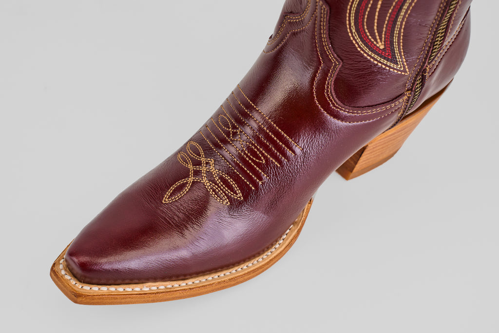 A close-up of the Allison Blackberry - Tall Shaft - Snip Toe boot, featuring a polished dark brown finish, decorative stitching, a pointed snip toe, and a light wooden heel, set against a plain light gray background.