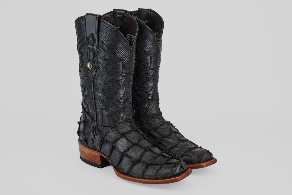 The Conor - Matte Black - Narrow Square Toe cowboy boots feature scale-like textured fish leather, detailed stitching, brown soles, and low wooden heels. They are shown on a light gray background.