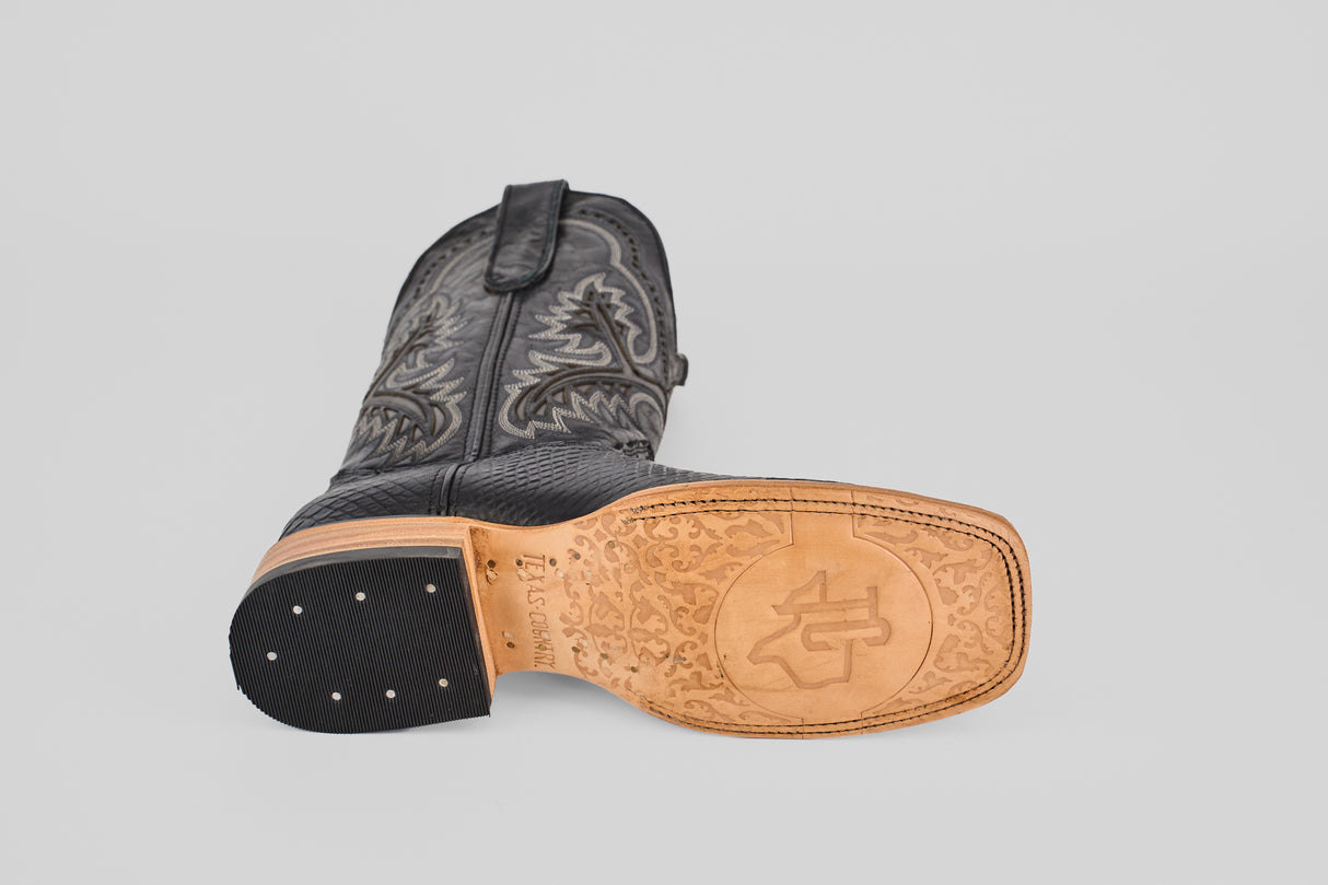 The Python Black - Embossed Leather - Square Toe (E425) men's boot lies on its side, showing a decorative stitched upper, tan leather sole with embossed designs, and small metal studs on the heel.