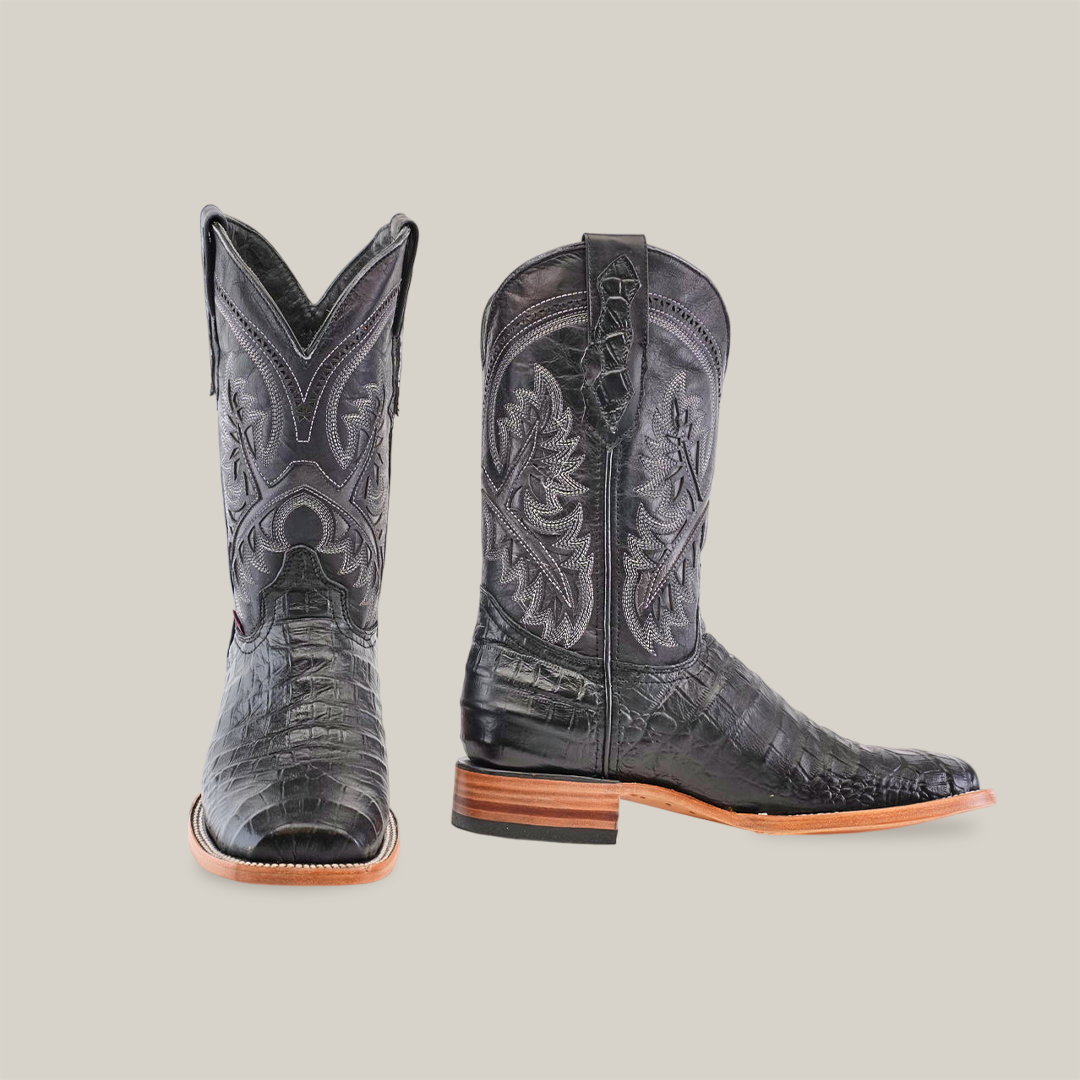 The Caiman Belly Print Black - Square Toe boots feature premium cowhide leather, intricate stitching, a sleek black design with Caiman Belly texture. One boot stands upright showing the toe, while the other is angled to highlight the heel and side pattern. The soles are light tan.