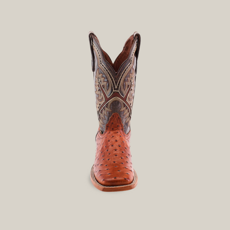 The Karoo Ostrich Print Cogñac - Square Toe is a handcrafted cowboy boot featuring a light brown ostrich print foot and a dark brown upper with intricate details. It stands upright on a white background, highlighting its pointed toe and decorative stitching.